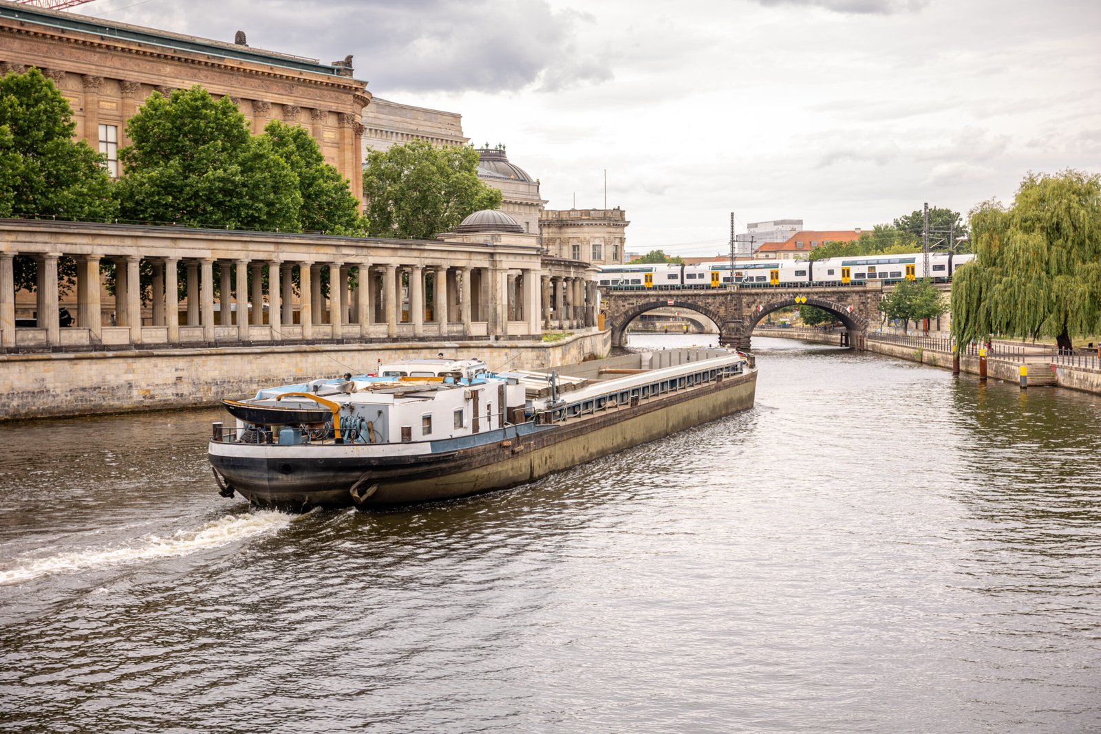 A freight vessel sails under a stone bridge while a train passes above. Classical architecture and intermodal transport meet in a scenic frame.