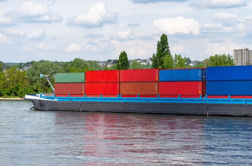 A barge carrying many containers on the Rhine in western Germany, trees and buildings in the background.